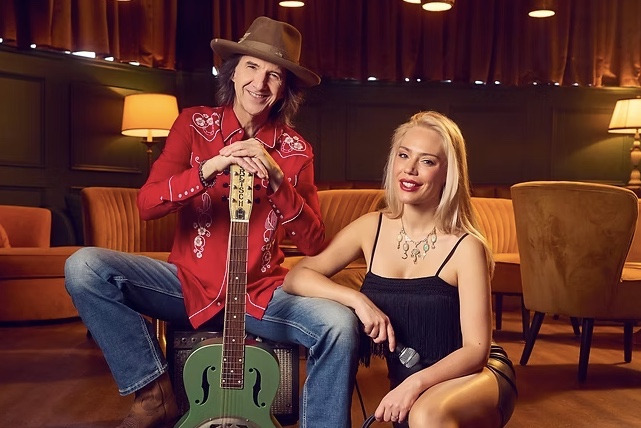 Dans un salon aux lumières tamisées, deux personnes posent près d’une guitare, l’une tenant un micro
